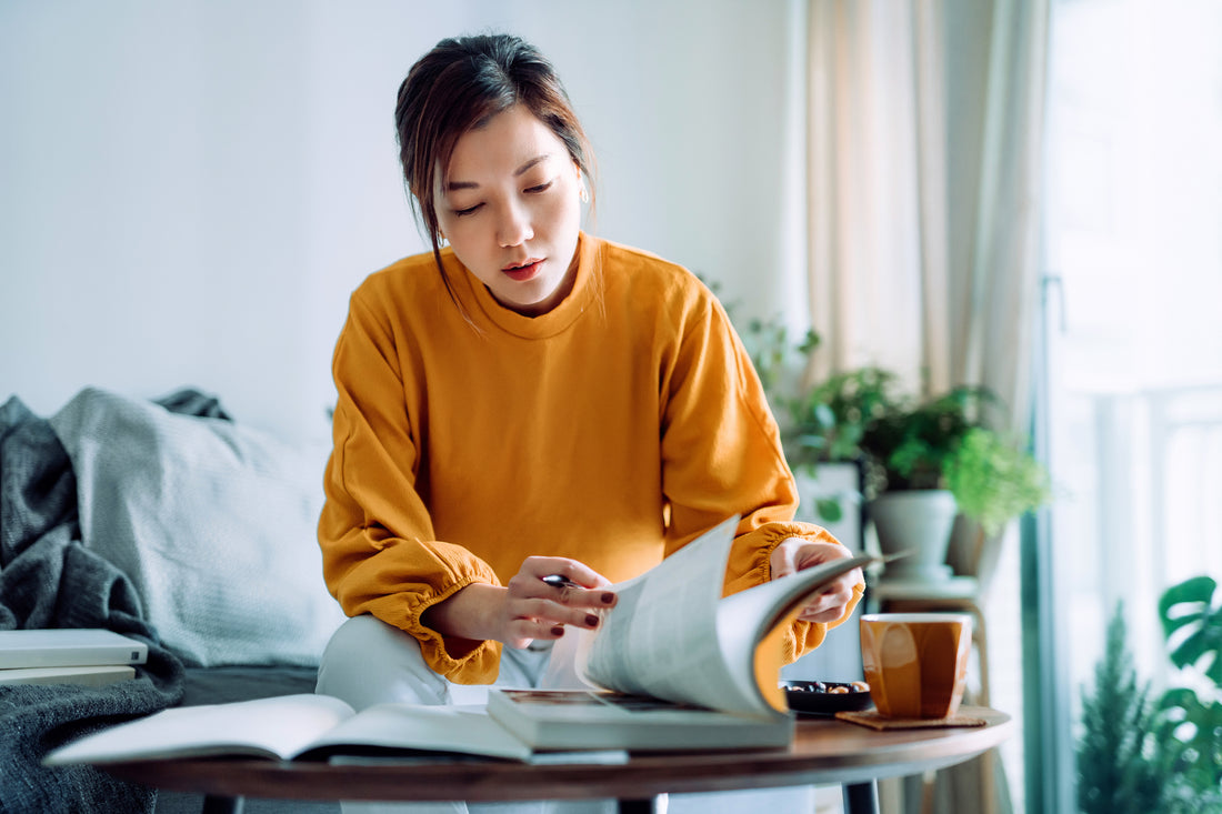 Focused young Asian woman reading a book and making notes at home, concentrating on her studies. 